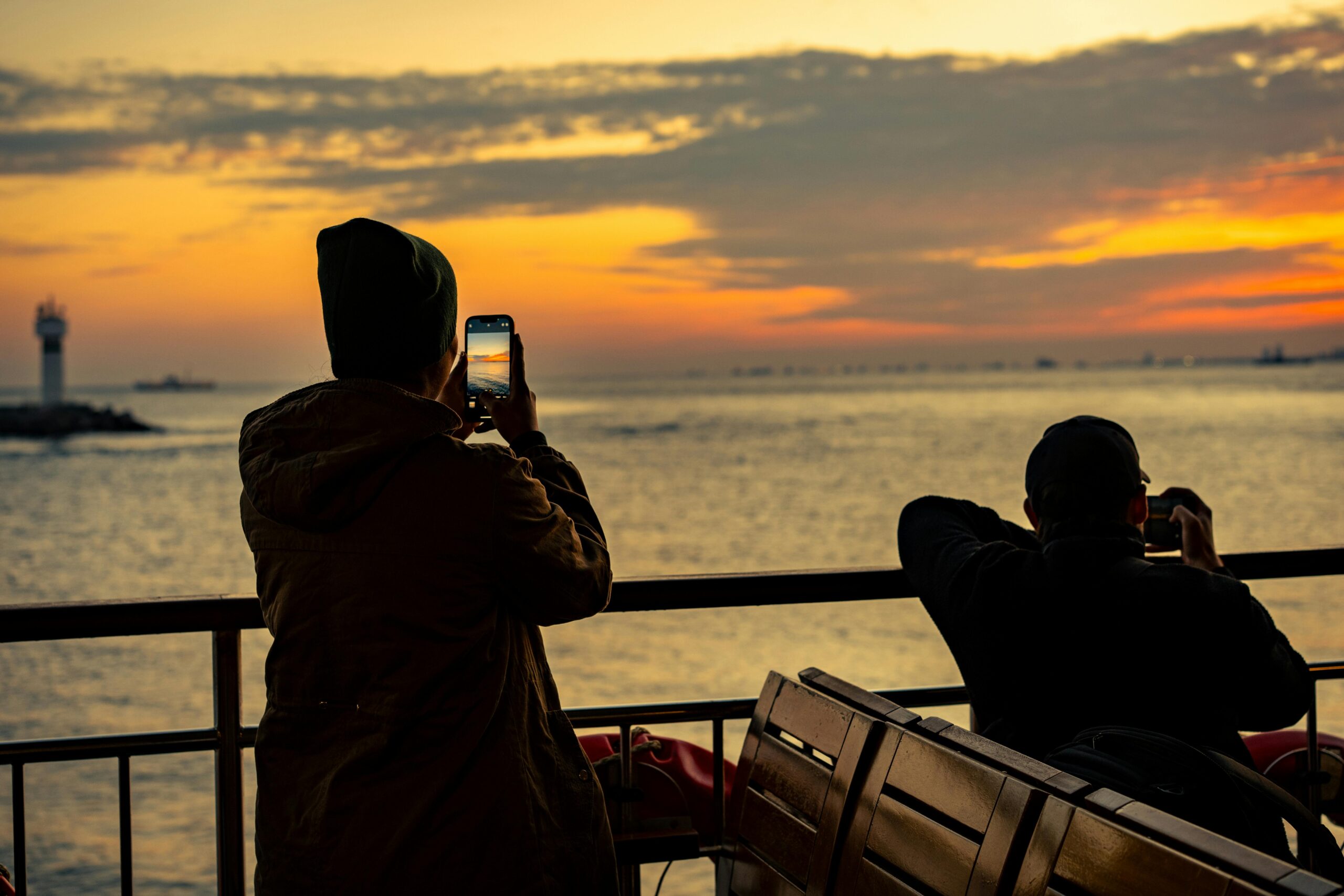 Two people photographing a stunning sunset by the sea, with a lighthouse in view.