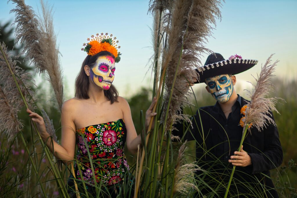 A couple celebrates Día de Muertos in traditional costumes with beautiful face paint and floral accents outdoors.