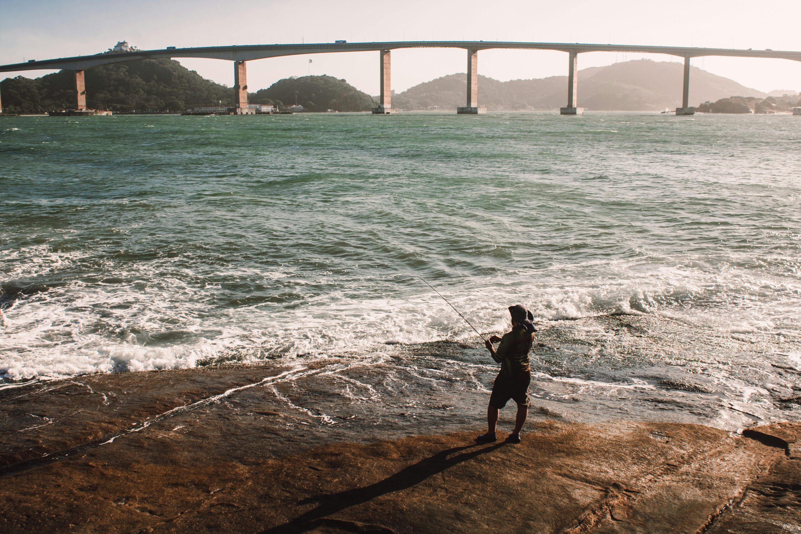 A person enjoys fishing by the coast with a scenic bridge in the background.