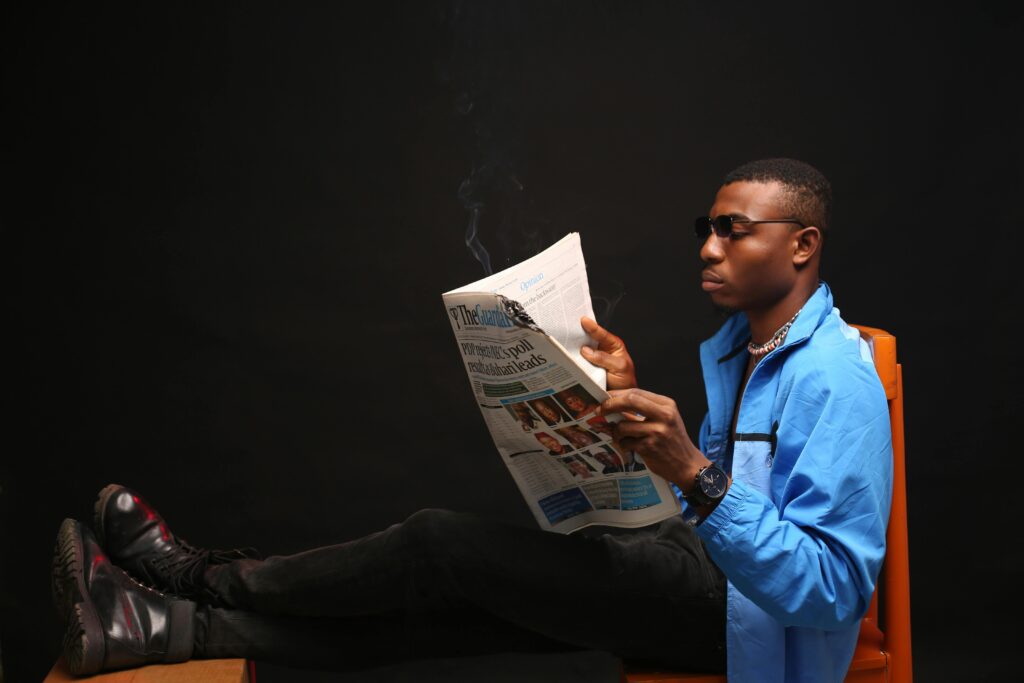 Adult male relaxing and reading newspaper indoors, dressed casually in a blue jacket and sunglasses.