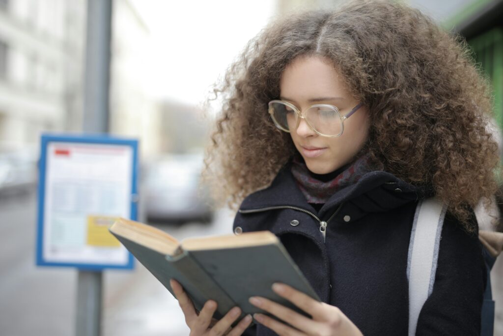 A young woman with glasses and curly hair reads a book while standing on a city street, captured in natural light.