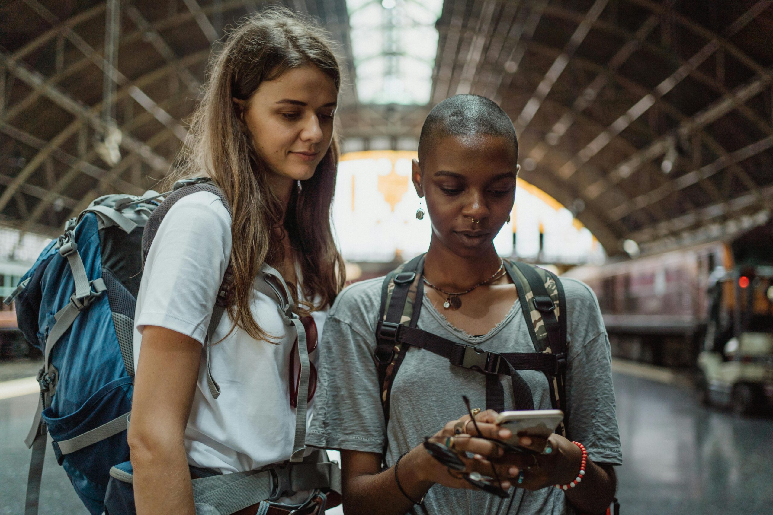 Two diverse female backpackers checking phone at a train station, ready to travel.