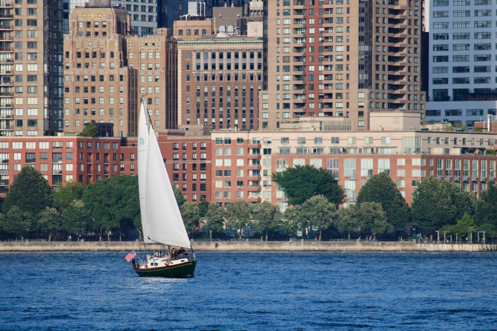 A sailboat glides along the Jersey City waterfront, with high-rise buildings in the background.