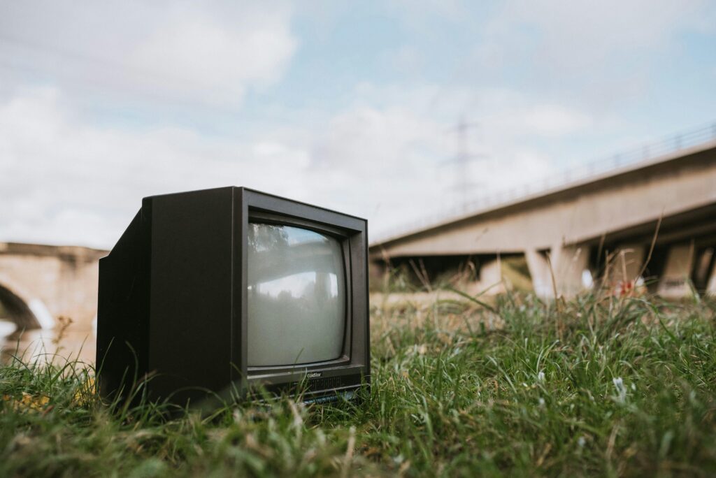 A retro television set placed in a grass field near a bridge, showcasing nostalgia and obsolescence.