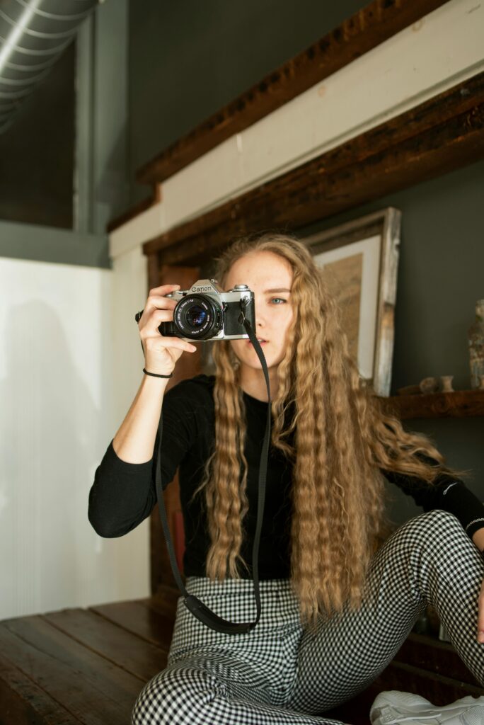 A young woman with long blonde hair takes a photo indoors, showcasing a relaxed lifestyle.