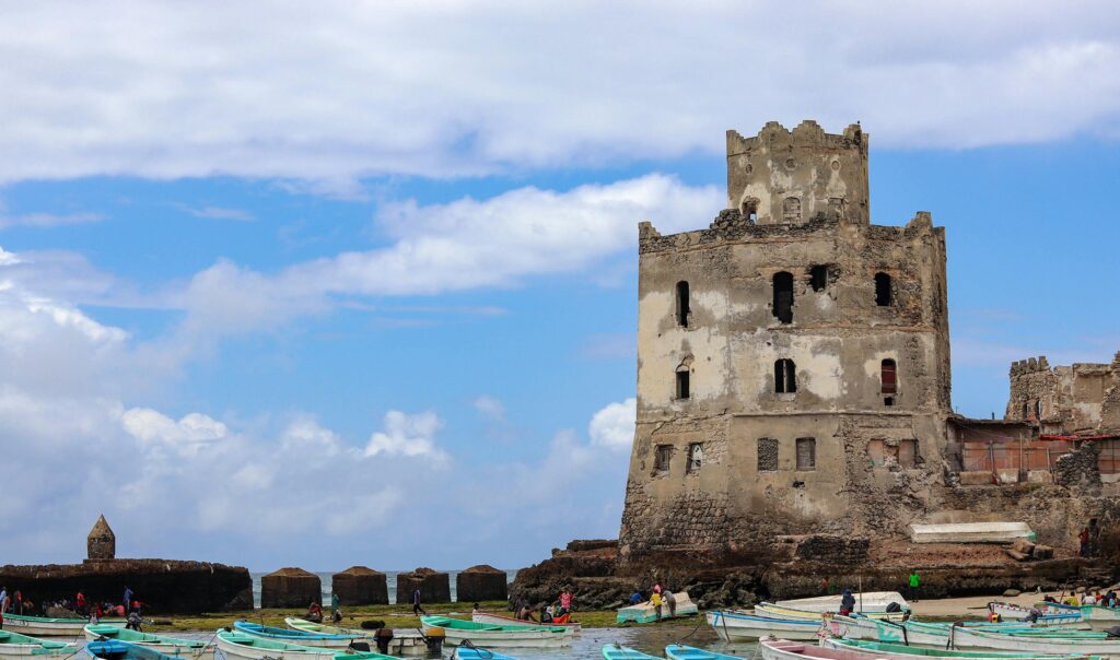 Explore the ancient coastal ruins of Mogadishu with colorful fishing boats in the foreground under a vibrant blue sky.