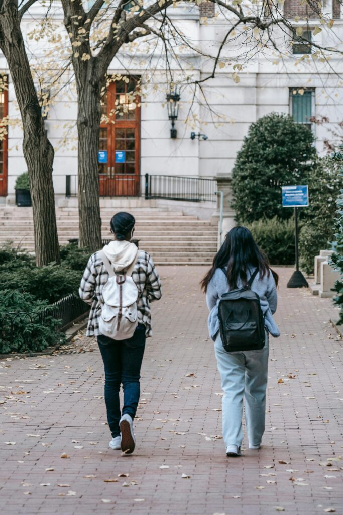Full body back view of anonymous female students in casual outfit with backpacks walking along pavement in university campus
