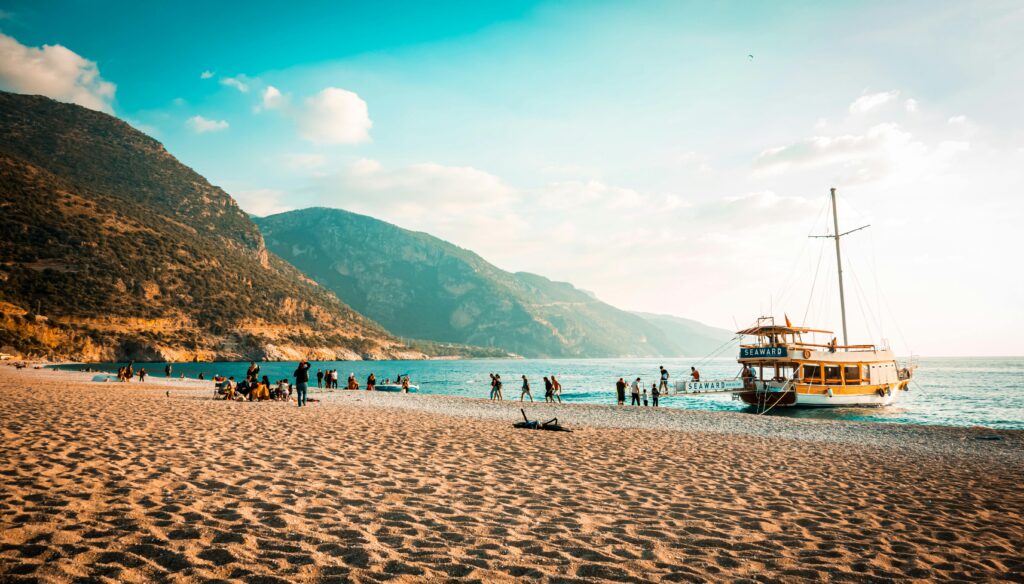 Tourists enjoying a beach with mountains and a boat named Seaward in view.