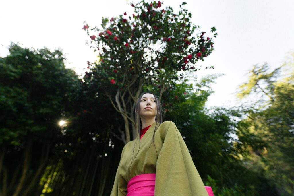 A woman in a kimono stands gracefully in a Japanese garden, surrounded by trees and spring blossoms.