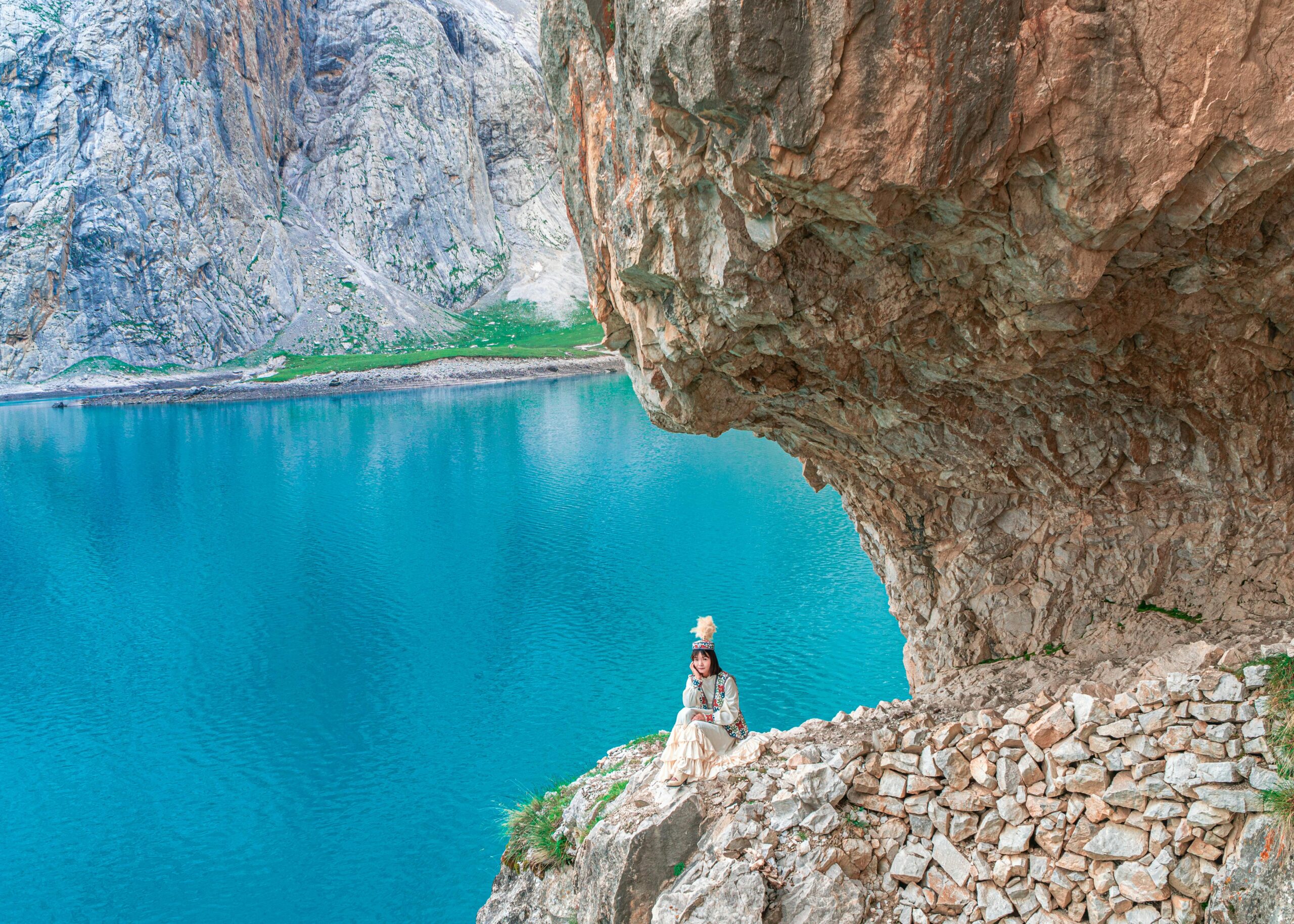 Woman in traditional attire sits by a turquoise lake in Xinjiang's dramatic cliffside terrain.