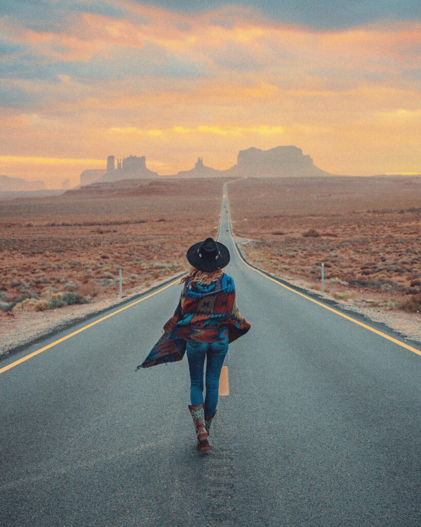 Woman walking on a desert road towards Monument Valley under a dramatic sunset sky.