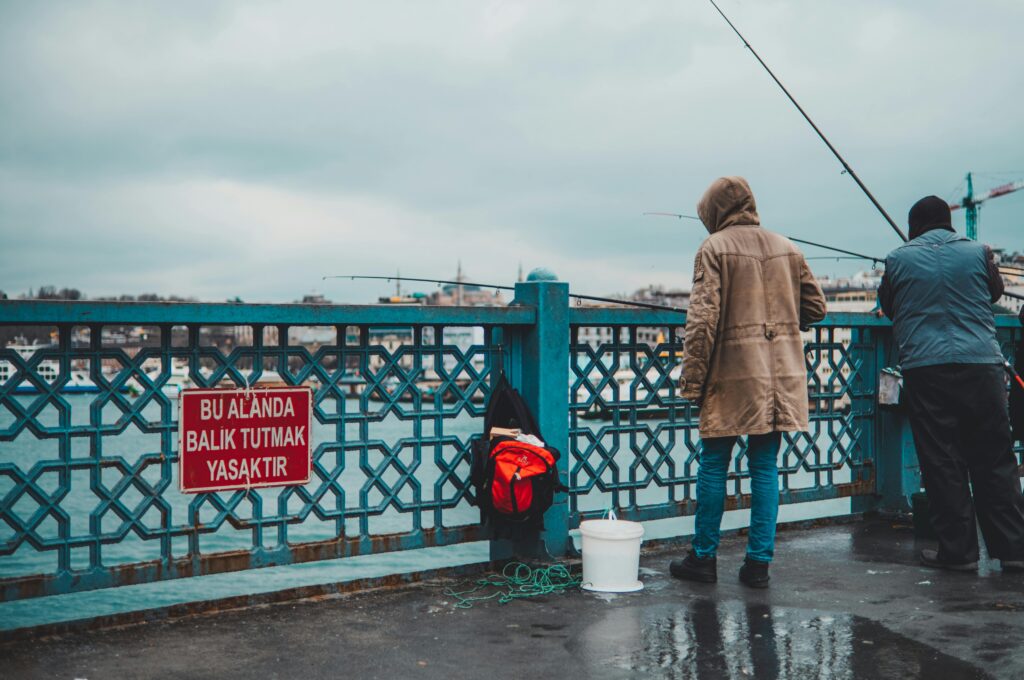 Two people fishing in front of a 'No Fishing' sign on a bridge in Istanbul.