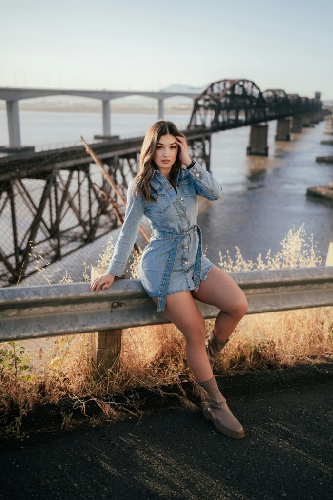 Fashionable woman poses by a scenic river bridge in daylight, showcasing a trendy denim outfit.