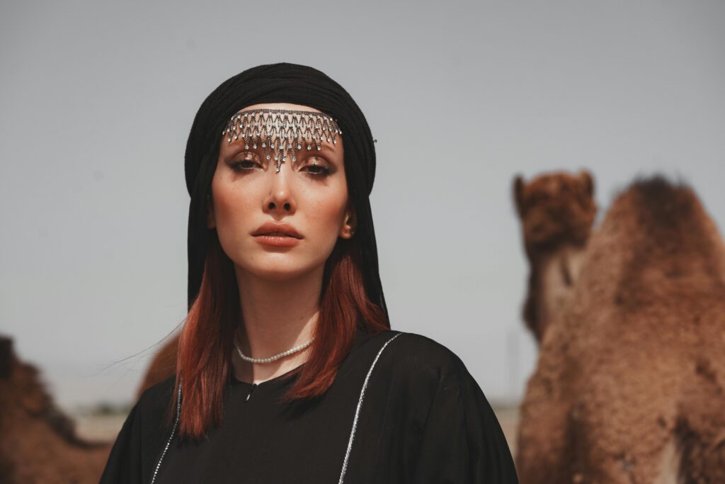A woman in traditional dress stands with camels in a desert setting.