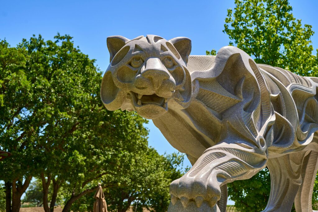 Artistic stone tiger sculpture set against lush green trees on a sunny day.