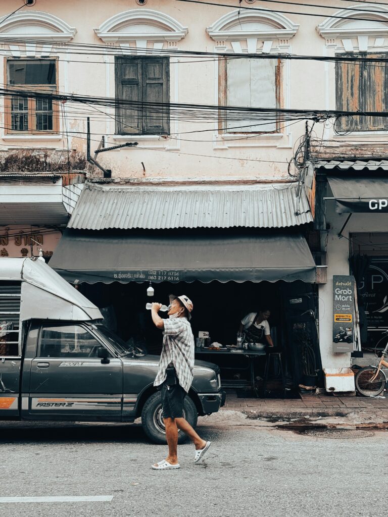 Man drinks water while walking past a food stall on a bustling Bangkok street.