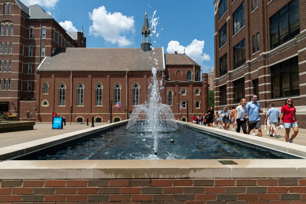 Daytime cityscape featuring a fountain, historic buildings, and pedestrians enjoying a sunny day.