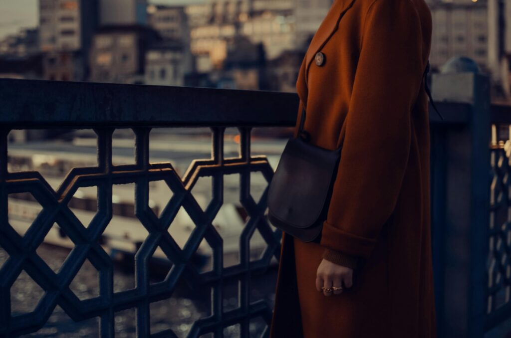Chic woman in a rust-colored coat standing on an urban bridge, showcasing city fashion style.