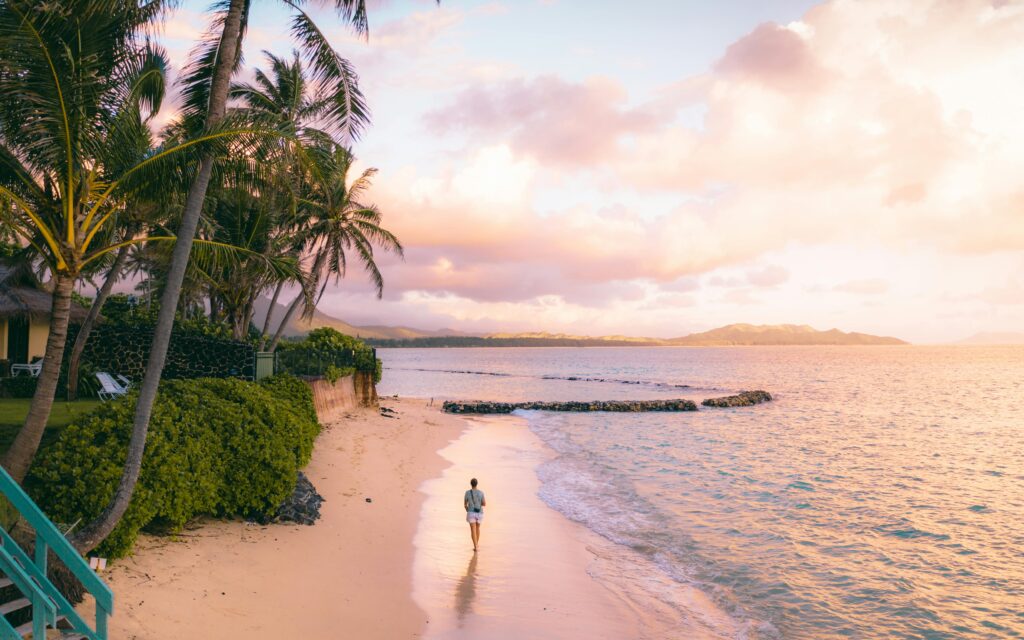 Person walks along Waimanalo Beach, Hawaii at sunrise with palm trees and golden sky.