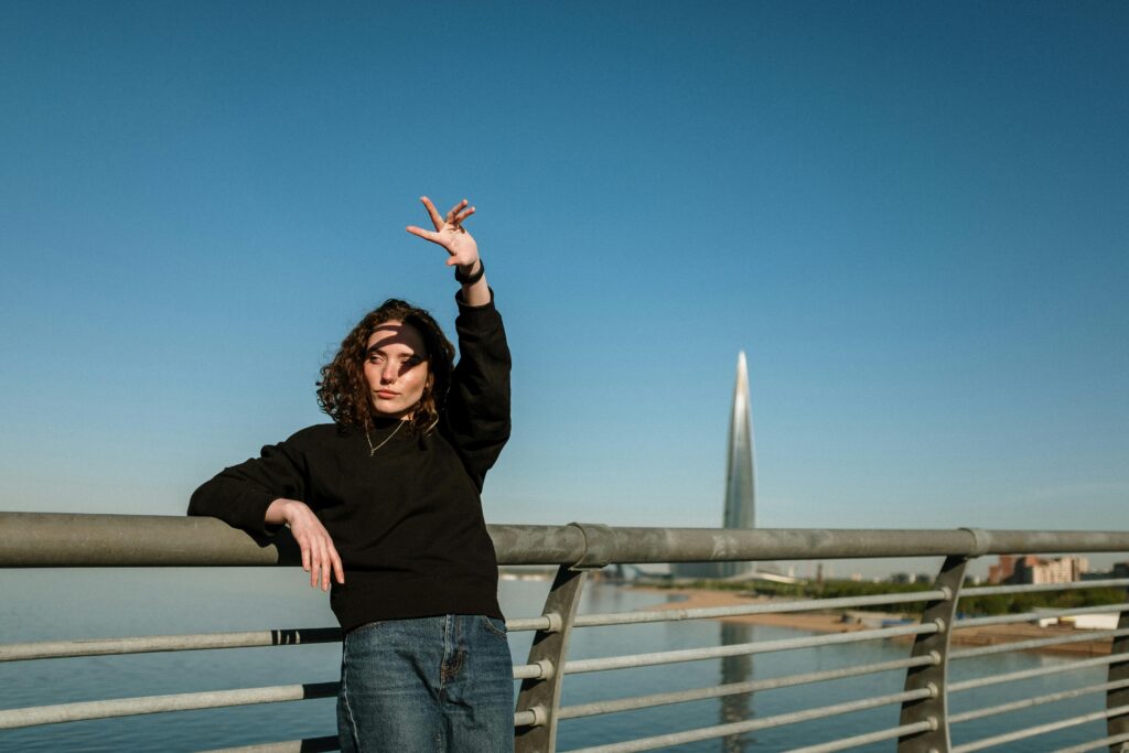 A young woman in casual attire poses on a bridge with a cityscape background featuring a modern skyscraper.