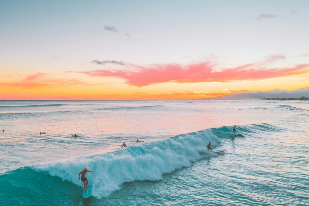 Stunning sunset view of surfers riding waves in Hawaii, showcasing beautiful ocean and sky colors.