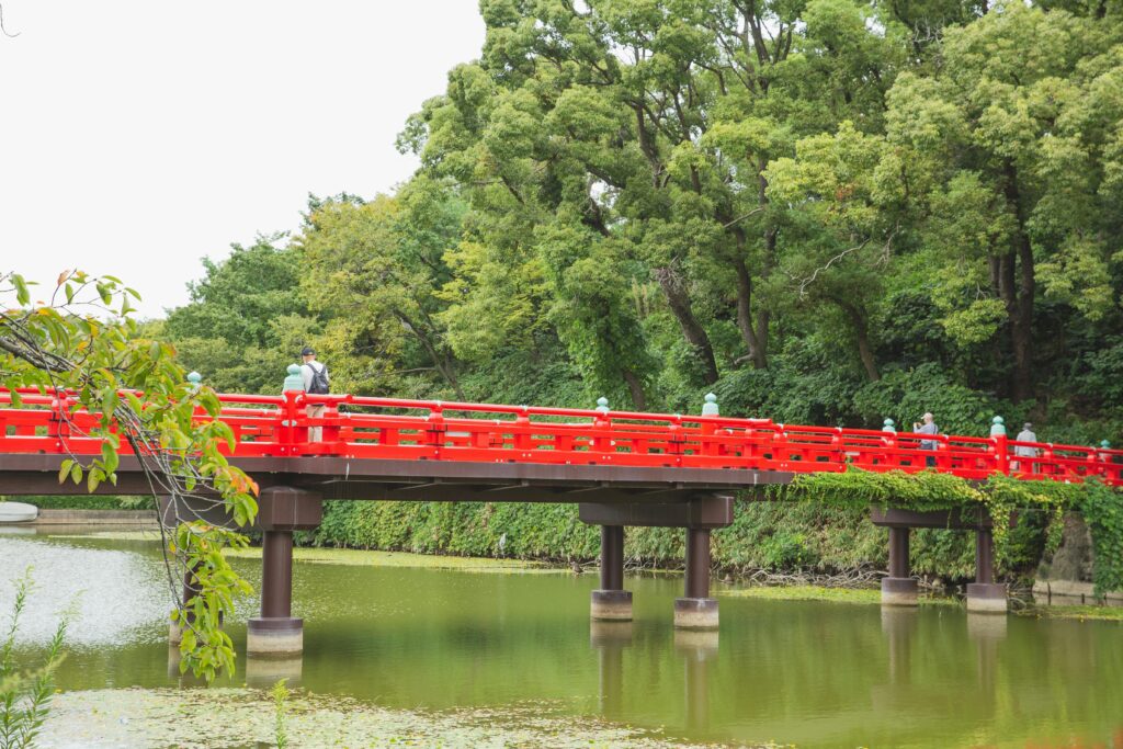 Tourists walking on red bridge crossing green pond located among lush tropical forest in Japan