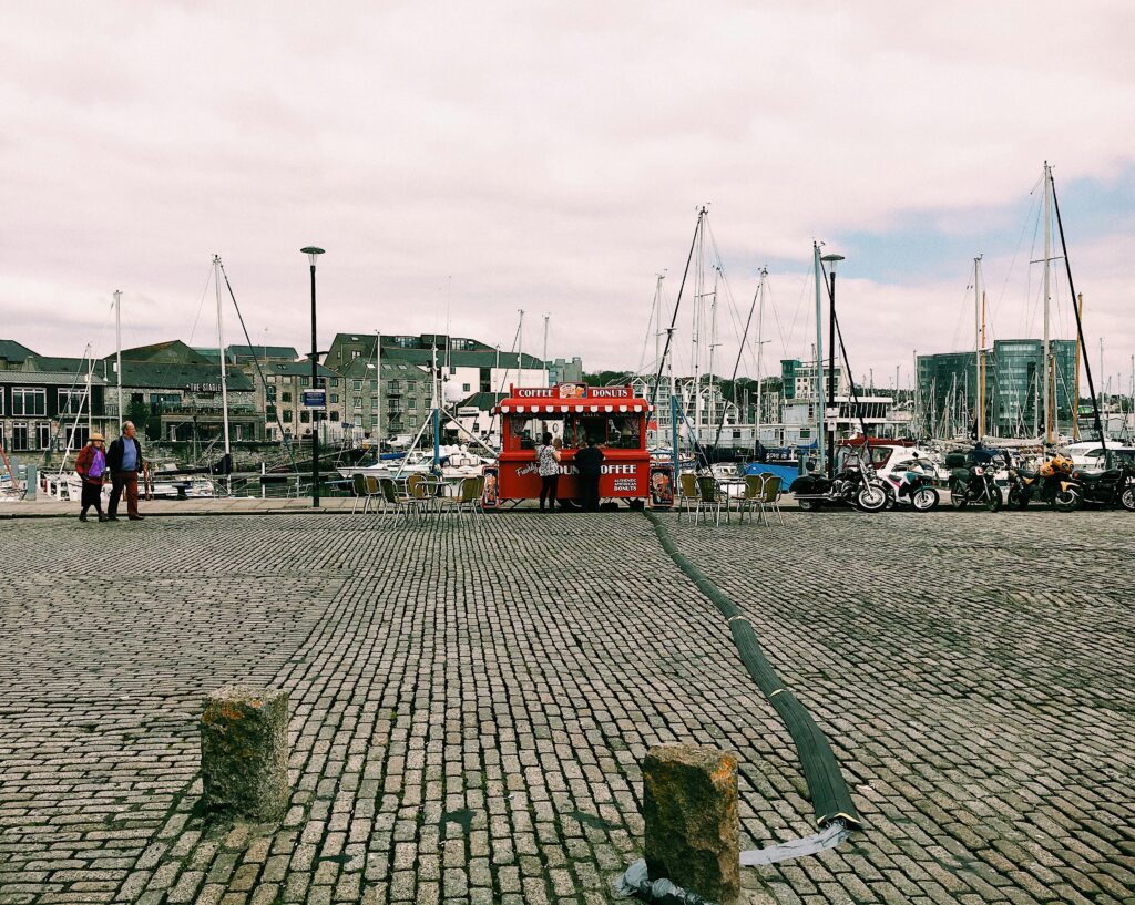 Modern harbor with people walking on embankment near street stall and motorcycles near river with moored boats and buildings on shore under cloudy sky in town in daylight