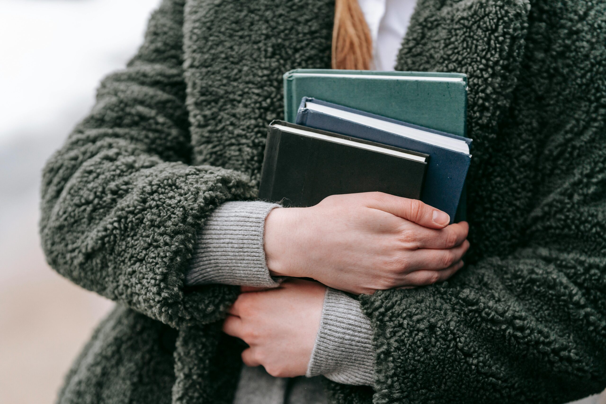 A woman embraces several hardcover books while wearing a warm, green coat on a chilly day.