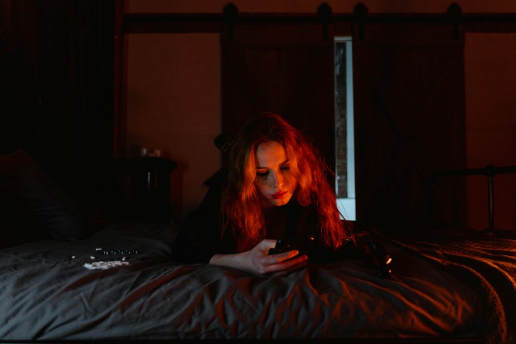 A young woman lying on a bed in a dark room, engrossed in her cellphone.