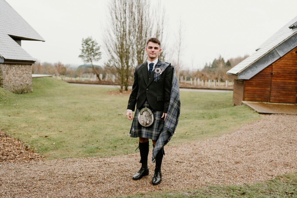 Young man in Scottish attire standing outside on a farm, ideal for wedding themes.