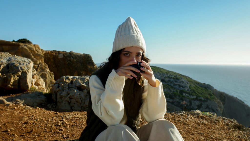 A woman in a cozy beanie drinks coffee by the ocean on a sunny day.