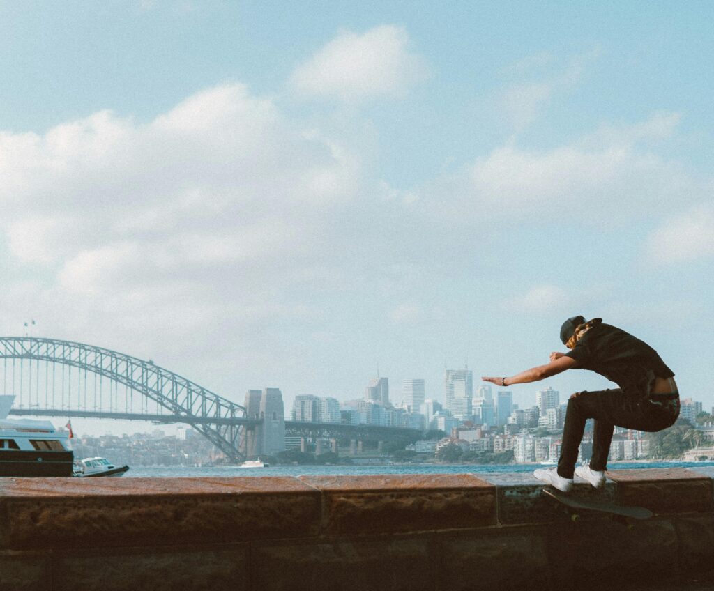A skateboarder performs a trick near the iconic Sydney Harbor Bridge on a sunny day.