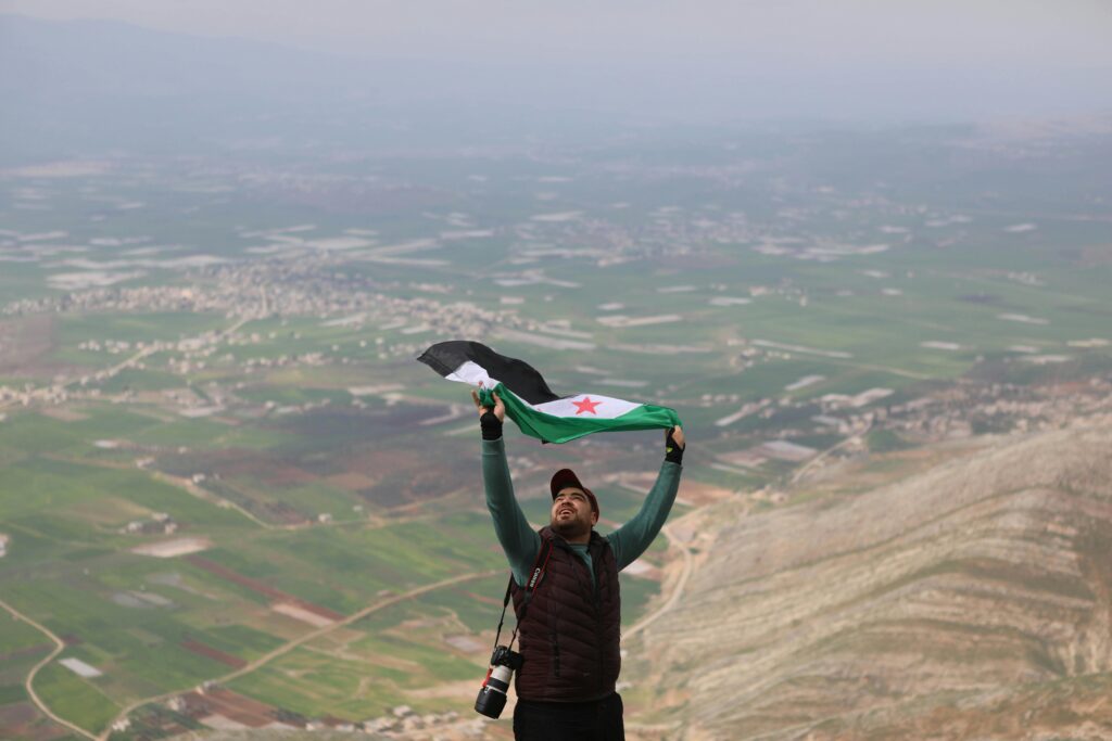 Man celebrates with Syrian flag atop a mountain overlooking the Idlib countryside.