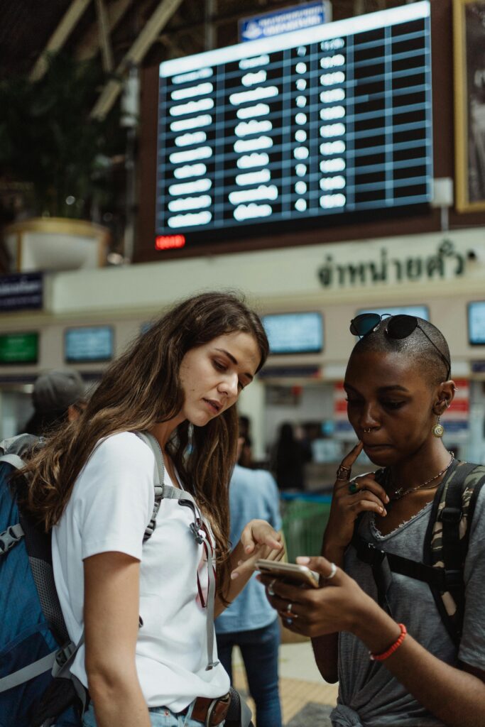 Two women with backpacks check a train timetable at a busy station.