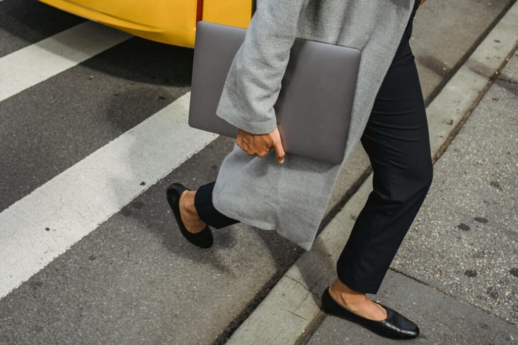 Crop anonymous female in casual outfit holding netbook while crossing asphalt road in city