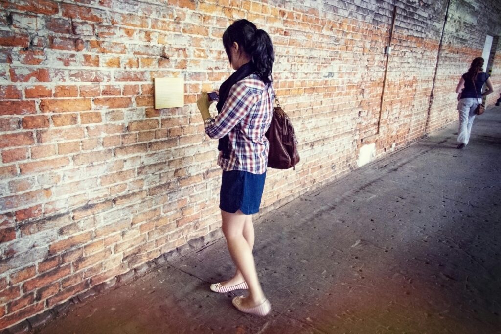 A young woman stands reading a plaque on an exposed brick wall in an industrial setting.