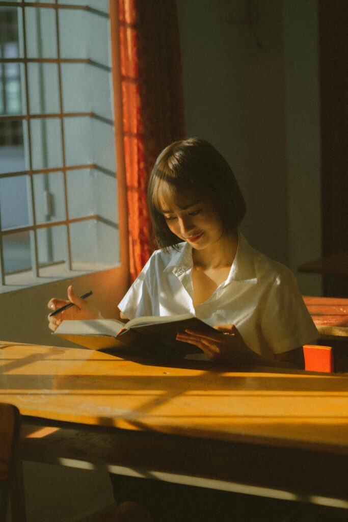 Asian woman in a school uniform reading at a sunlit desk indoors, capturing a serene moment of learning.