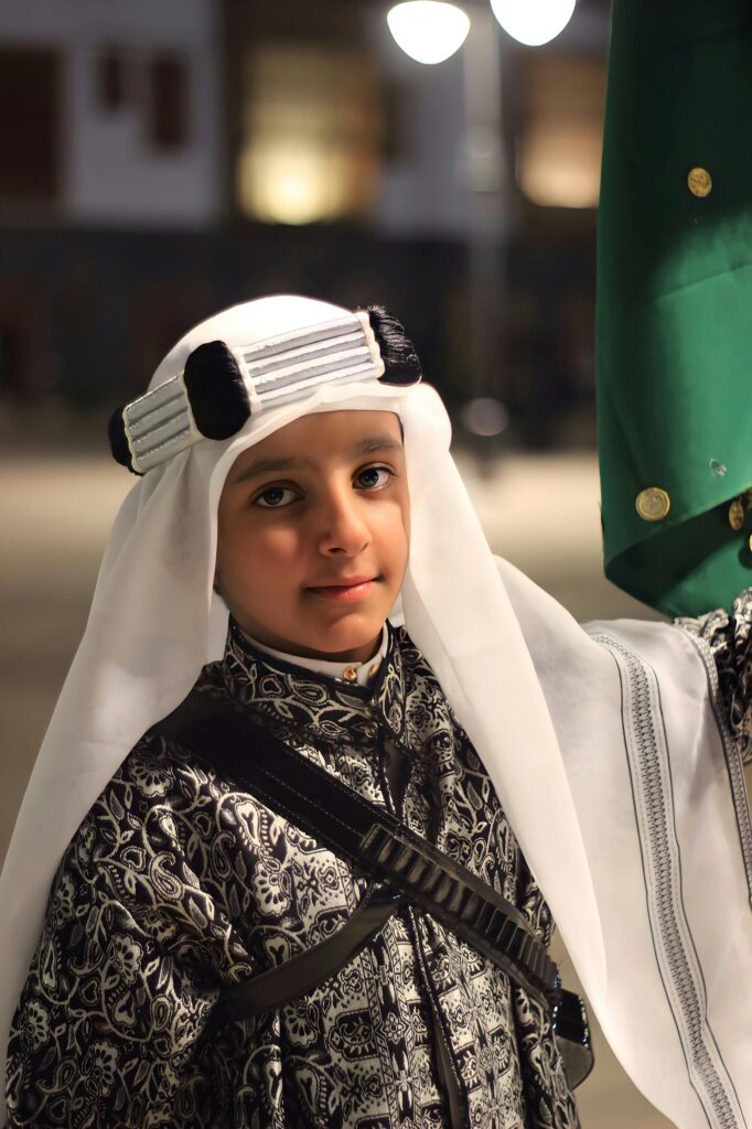 A young Saudi boy poses in traditional attire against a street backdrop at night in Saudi Arabia.