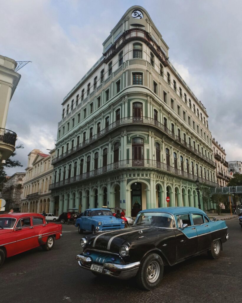 Vintage cars drive past the iconic Hotel Saratoga in Havana, Cuba, capturing a retro urban vibe.