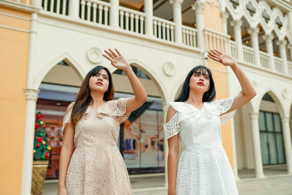 Two women in elegant dresses posing gracefully outdoors against an architectural backdrop.