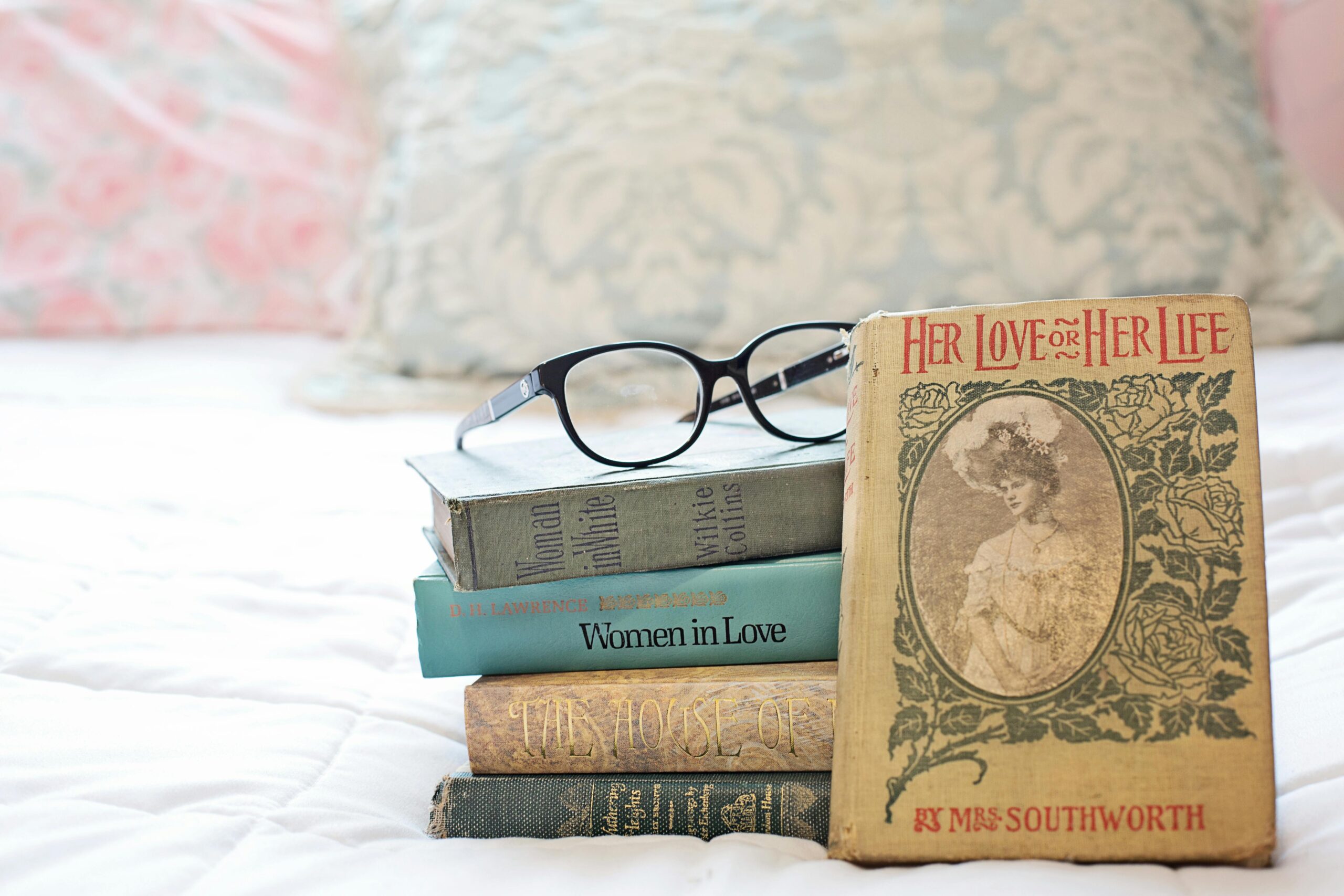 A stack of vintage romance novels with eyeglasses on a white bed.