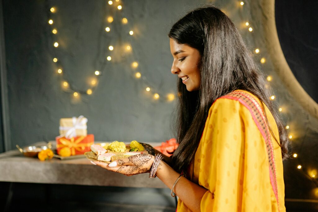 Side view of a woman holding traditional Diwali sweets with festive lights in the background.