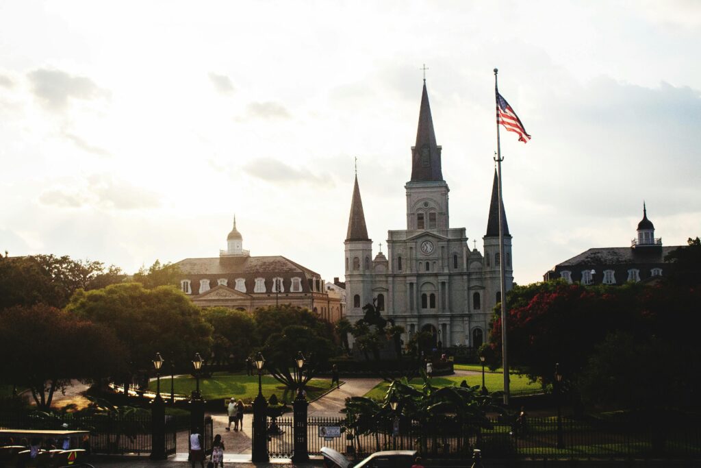 St. Louis Cathedral in New Orleans with an American flag, trees, and a garden at dusk. Iconic religious landmark.