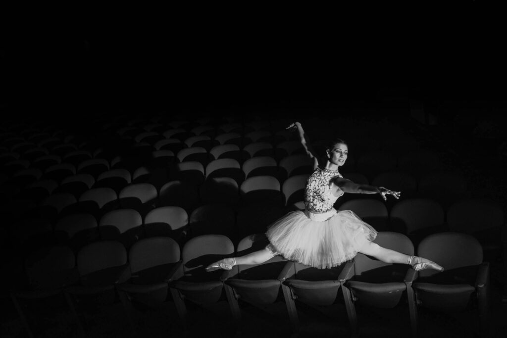 Ballet dancer gracefully leaps amid empty theater seats, captured in black and white.