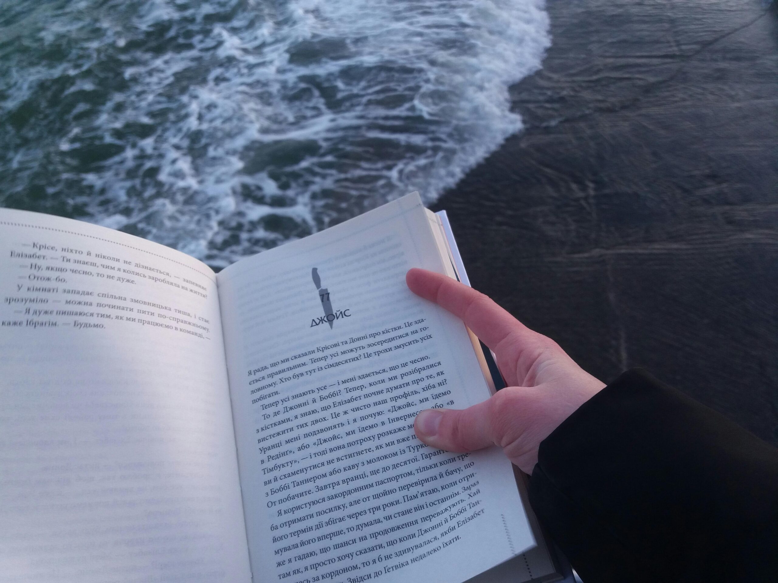 A person holds and reads a book by the ocean, enjoying a peaceful moment by the water.