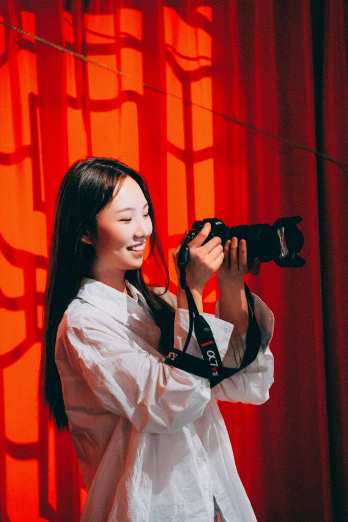 Portrait of a smiling young woman photographer with a camera indoors against red curtains.