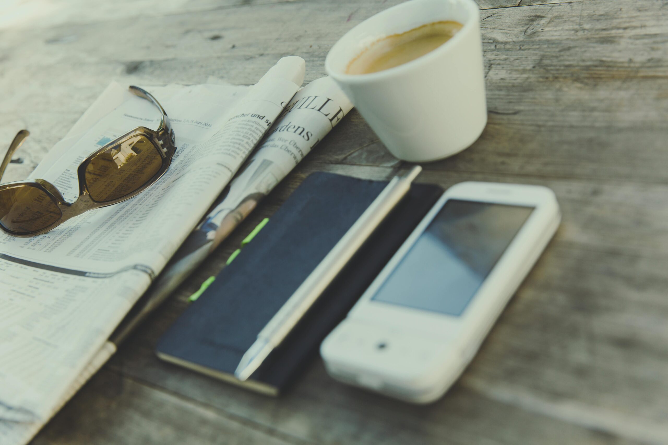 Relaxing scene featuring coffee, newspaper, sunglasses, notebook, and smartphone on a rustic wooden table.