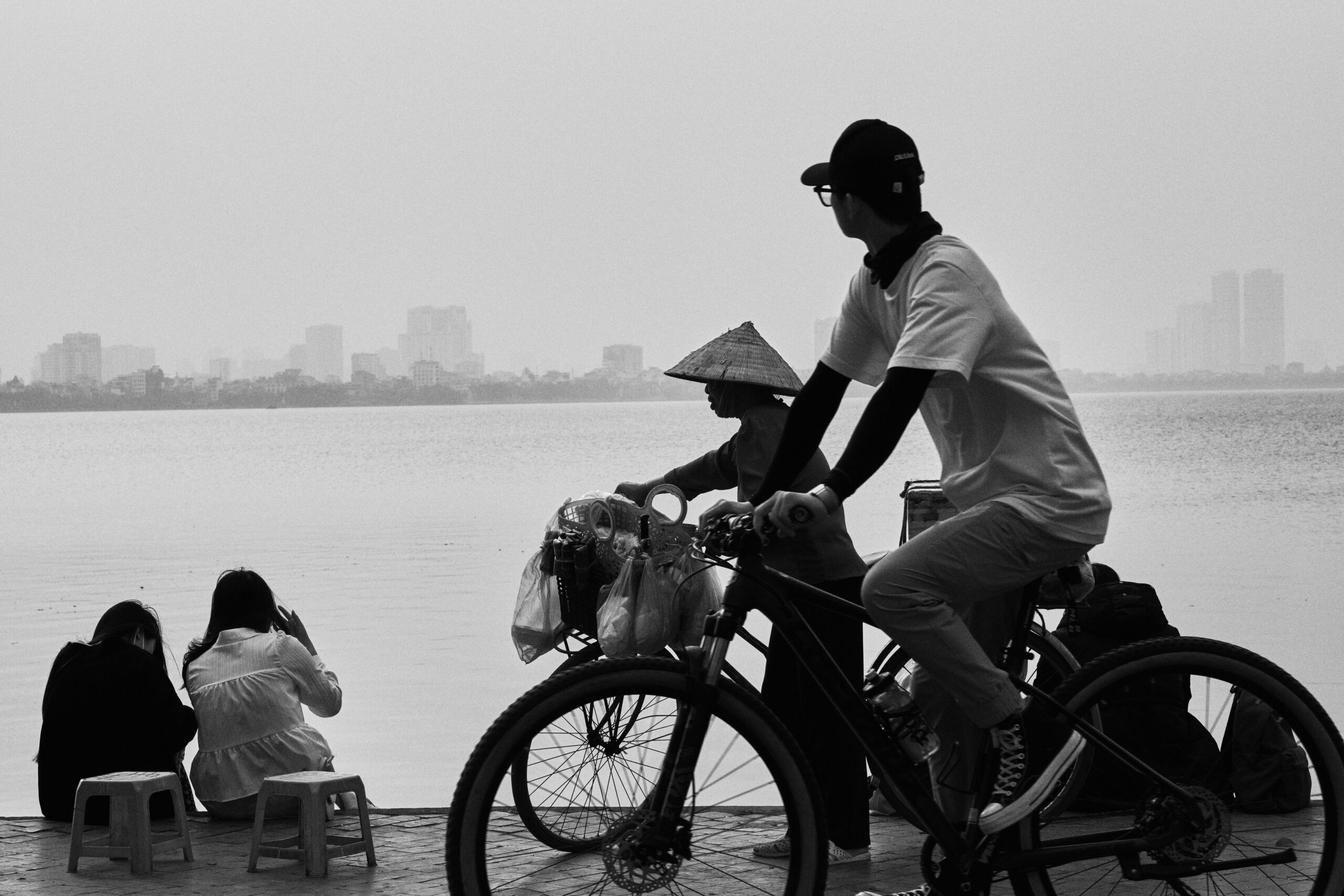 A serene scene of daily life by a lake in Hanoi, capturing locals relaxing, biking, and enjoying the view.