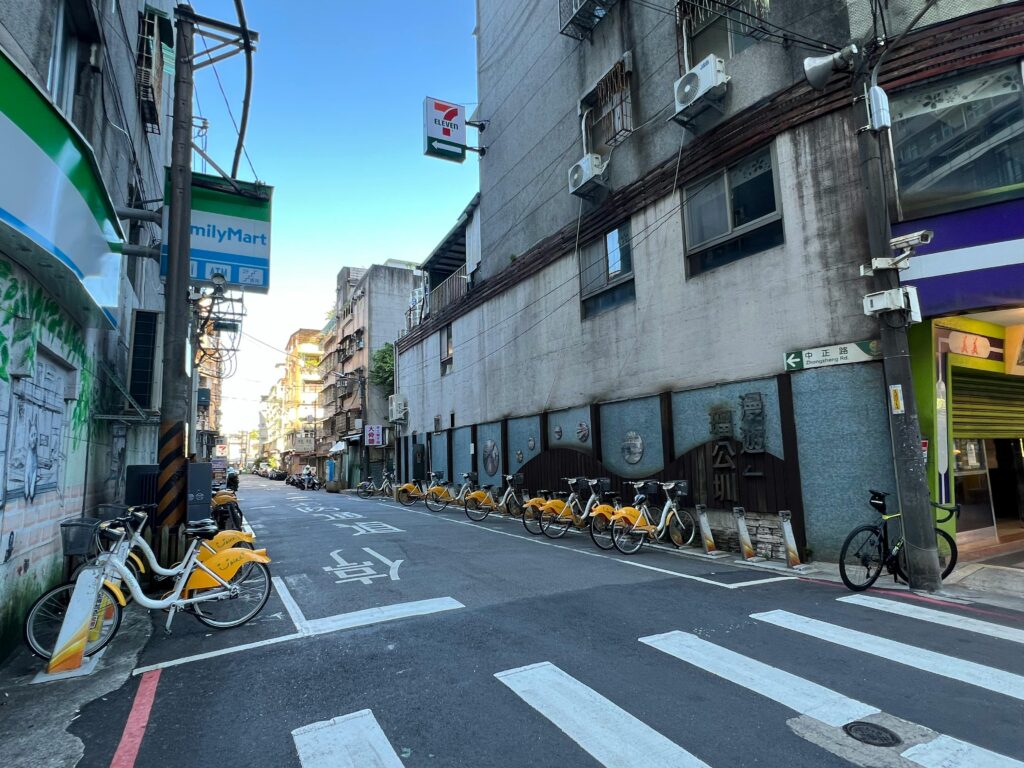 Urban street scene in New Taipei City featuring parked yellow bicycles and local shops under a clear sky.