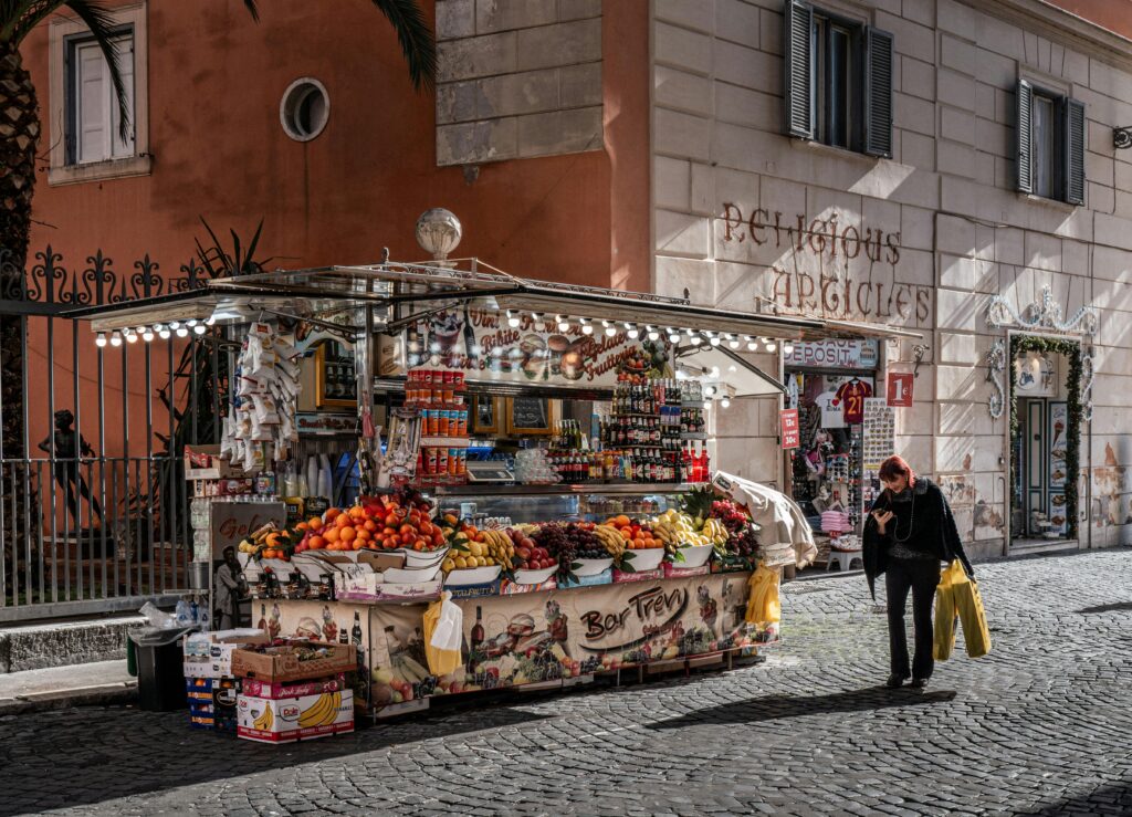 Colorful street market stall selling fruits and drinks in Rome city center with a passerby.